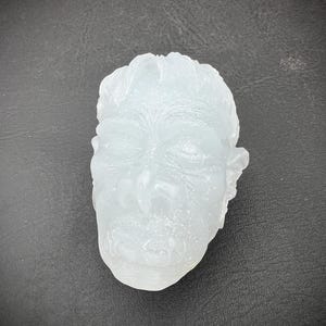 Può includere: Un volto scolpito traslucido azzurro con occhi chiusi, naso prominente e lineamenti dettagliati. La scultura ha una superficie strutturata su uno sfondo scuro.