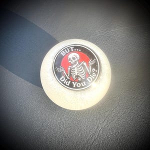 May include: A round, off-white decorative object with a graphic of a skeleton inside a black circle with red background. The text reads "BUT... Did You Die?"