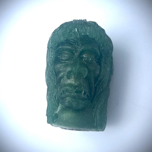 May include: A green, carved sculpture of a face with closed eyes and a stitched scar. The face has a beard and long hair. The sculpture is set against a white background.