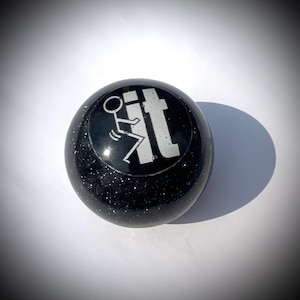 May include: A black, spherical object with a glittery texture. The object features a white graphic of a stick figure kicking the word "it". The object is likely a decorative item or novelty product.