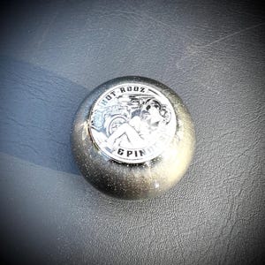 May include: A silver and glittery shift knob with the text "HOT RODZ & PINUPS" around a graphic of a classic car and a woman. The knob is round and has a metallic sheen.