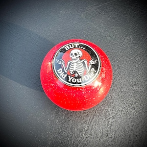May include: A red, round button with a glittery finish. The button features a black and white graphic of a skeleton with the text "BUT... Did You Die?" in a circular design.