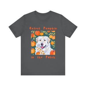 Puede incluir: Camiseta gris oscuro con un diseño temático de calabaza que presenta un perro blanco sonriente. El texto "Cutest Pumpkin in the Patch" está impreso encima y debajo de la ilustración del perro, rodeado de calabazas naranjas y hojas verdes.