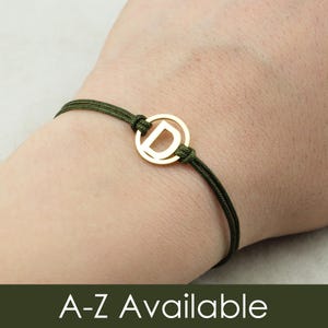 Könnte beinhalten: Ein Armband mit einer dunkelolivgrünen Kordel und einem goldfarbenen, kreisförmigen Anhänger, in dessen Mitte der Buchstabe D eingearbeitet ist. Die Kordel ist an beiden Seiten des Anhängers verknotet und bildet eine durchgehende Schlaufe.