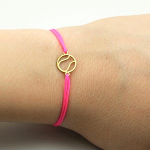 Könnte beinhalten: Ein Armband mit einem goldfarbenen Tennisball-Anhänger, der an einem leuchtend pinkfarbenen Band befestigt ist. Der Anhänger besteht aus einer kreisförmigen Kontur mit den typischen geschwungenen Linien eines Tennisballs. Das Band ist dünn.