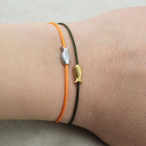 Può includere: Due braccialetti con ciondoli a forma di pesce. Uno ha un cordino arancione e un ciondolo a forma di pesce argentato. L'altro ha un cordino verde scuro e un ciondolo a forma di pesce dorato. Design semplice e minimalista.