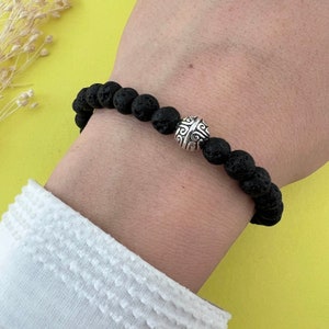 Könnte beinhalten: Ein schwarzes Armband aus Lavasteinen mit einem silbernen Perlenanhänger mit dekorativem Design. Das Armband wird am Handgelenk getragen.