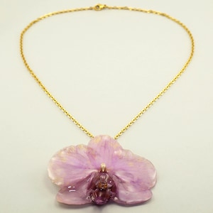 Könnte beinhalten: Eine goldfarbene Halskette mit einem Anhänger in Form einer hellvioletten Orchideenblüte. Die Orchidee hat ein detailliertes, realistisches Design mit einem dunkleren violetten Zentrum und goldenen Akzenten.