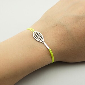 Op de afbeelding: Een armband met een fel neongeel koord en een zilverkleurige tennisracketbedel. De bedel heeft een gedetailleerd rasterpatroon dat de snaren van het racket voorstelt en is stevig bevestigd aan het levendige koord rond de pols.