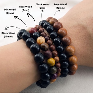 May include: Wooden bead bracelets in a range of colours and sizes. The bracelets feature black wood (10mm and 8mm), rose wood (10mm and 6mm), and mix wood (8mm) beads. The image displays the bracelets on a wrist.