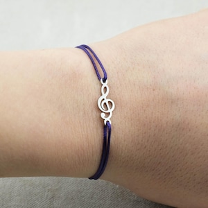 Könnte beinhalten: Ein Armband mit einem silberfarbenen Violinschlüssel-Anhänger, der mittig an einer doppelsträngigen violetten Kordel befestigt ist. Das metallische Musiksymbol ist oben und unten mit der dünnen, glatten violetten Schnur verbunden.