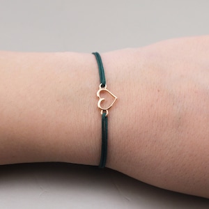 Può includere: Un braccialetto delicato caratterizzato da un sottile cordino verde scuro e un piccolo ciondolo in metallo dorato a forma di cuore. Il cuore è aperto al centro ed è collegato al cordino sia nella parte superiore che in quella inferiore.