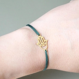 Könnte beinhalten: Ein Armband mit einem goldfarbenen Hamsa-Hand-Anhänger und einer dunkelgrünen Kordel. Die Hamsa-Hand ist ein Symbol für Schutz und Glück. Das Armband ist ein schlichtes, elegantes Schmuckstück.