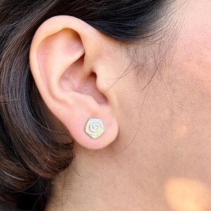 Könnte beinhalten: Nahaufnahme eines silbernen Ohrsteckers in Form einer Rose. Der Ohrring wird an einem Ohr getragen. Das Rosendesign hat eine spiralförmige Mitte und ist von Blütenblättern umgeben. Der Ohrring hat einen dezenten, metallischen Glanz.