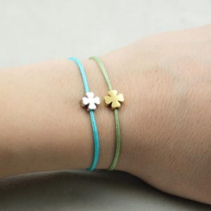 Könnte beinhalten: Zwei zarte Armbänder mit Kleeblatt-Anhängern. Eines hat ein silbernes Kleeblatt an einem türkisfarbenen Band, das andere ein goldenes Kleeblatt an einem grünen Band. Die Armbänder sind schlicht und elegant.