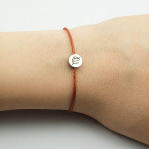 Könnte beinhalten: Ein dünnes Armband aus orangefarbener Kordel mit einem kleinen, runden, silberfarbenen Metallanhänger. Der Anhänger ist mit dem Tierkreiszeichen Jungfrau graviert, das wie ein stilisierter Buchstabe M mit einer Schlaufe am Ende aussieht.
