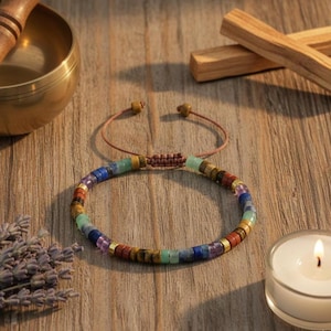 Può includere: Un braccialetto colorato con perline e una varietà di pietre preziose, tra cui pietre blu, verdi e rosse, è esposto su una superficie di legno. Il braccialetto è infilato su un cordoncino marrone e presenta distanziatori dorati. È un gioiello.