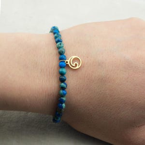 Può includere: Braccialetto di perline blu e turchese con un ciondolo dorato a forma di spirale. Il braccialetto è indossato su un polso. Le perline hanno un diametro di circa 0,6 cm.