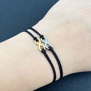 Pode incluir: Duas pulseiras com símbolo de infinito em um pulso. Uma pulseira é dourada e a outra é prateada. Ambas as pulseiras estão em um cordão preto.