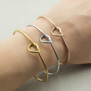 Puede incluir: Tres pulseras finas en oro, plata y oro rosa se muestran en una muñeca. Cada pulsera presenta un diseño de nudo en el centro. Las pulseras son sencillas y elegantes, adecuadas para el uso diario. Son de metal.