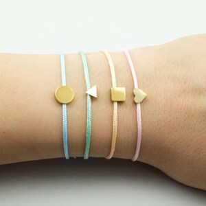 Könnte beinhalten: Vier zarte Armbänder mit goldfarbenen Metallanhängern in Form eines Kreises, eines Dreiecks, eines Quadrats und eines Herzens. Jeder Anhänger ist auf einem dünnen, gewebten Band in Pastelltönen wie Hellblau, Mintgrün, Blassgelb und Zartrosa zentriert.