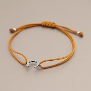 Könnte beinhalten: Ein senfgelbes Kordelarmband mit einem silberfarbenen Hufeisen-Anhänger aus Metall in der Mitte. Das Armband verfügt über einen verstellbaren Schiebeknoten-Verschluss mit zwei kleinen silberfarbenen Perlen an den Enden der Kordel für eine individuelle Passform.