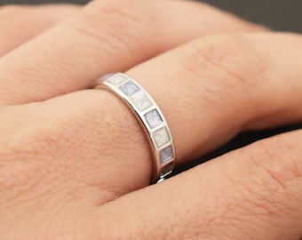 May include: A silver band ring with a row of square-shaped inlays. The inlays are a mix of white and light blue.
