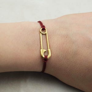 Könnte beinhalten: Ein Armband mit einer goldfarbenen Sicherheitsnadel und einer weinroten Kordel. Die Sicherheitsnadel ist der Blickfang, mit einem schlichten, eleganten Design. Das Armband wird am Handgelenk getragen und zeigt den Stil des Schmucks.