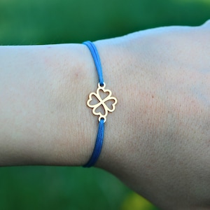 May include: A gold-coloured four-leaf clover charm bracelet with a bright blue cord. The clover is a classic symbol of luck and is centered on the bracelet. The bracelet is a simple, elegant design.