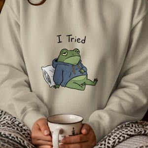 May include: Cream-colored sweatshirt with a cartoon frog wearing a blue hoodie and resting on a pillow. The text "I Tried" is above the frog. A white mug with a black design is held in the hands.