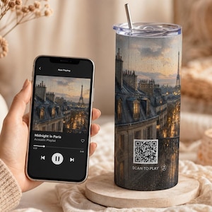 May include: A travel tumbler with a Parisian cityscape design, featuring the Eiffel Tower and streetlights. A smartphone displays a music playlist titled "Midnight in Paris." The tumbler has a QR code that says "SCAN TO PLAY."