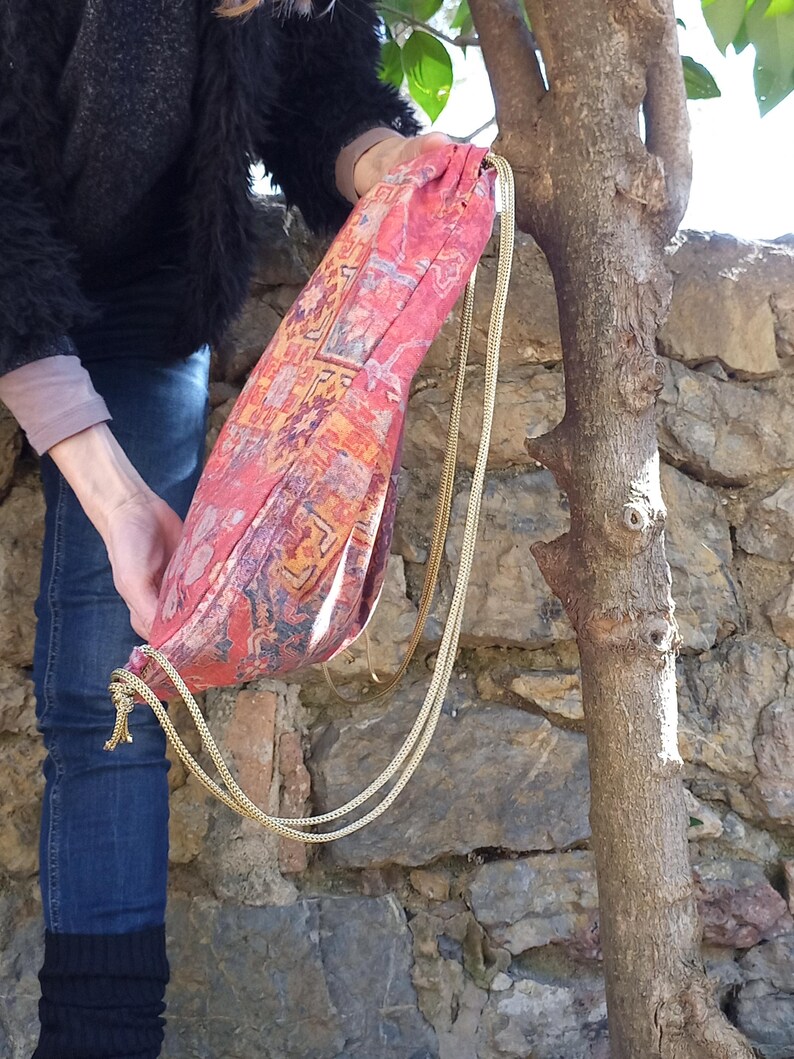 May include: A red and gold drawstring bag with a patterned design. The bag is made of fabric and has a long gold chain strap.