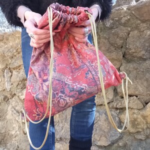 May include: A red and gold drawstring bag with a patterned design. The bag is made of fabric and has a gold chain drawstring.