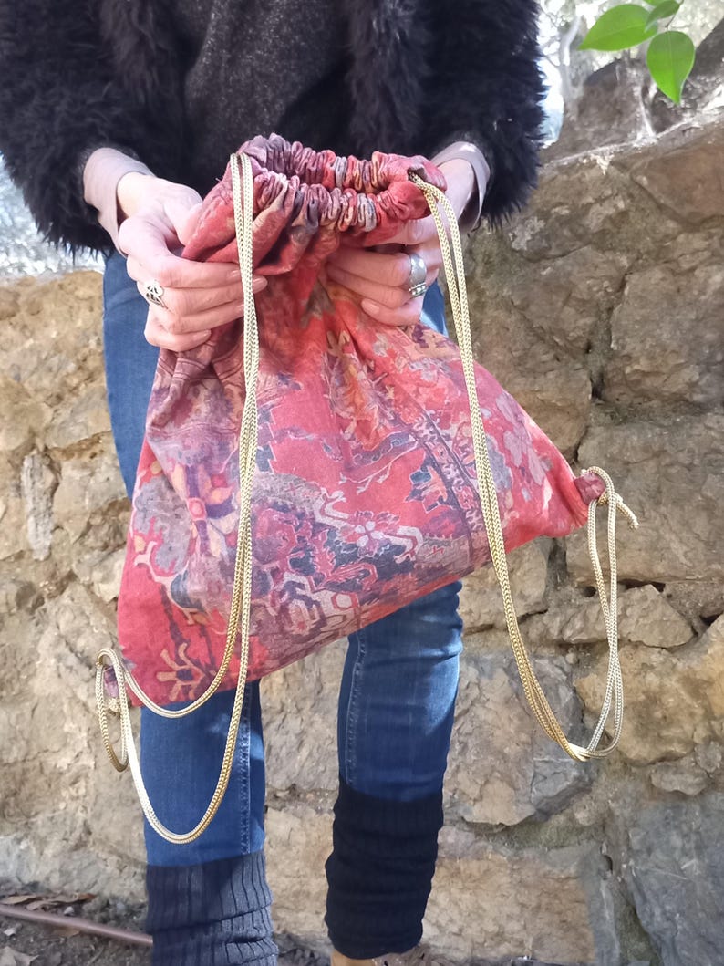 May include: A red and gold drawstring bag with a patterned design. The bag is made of fabric and has a gold chain drawstring.