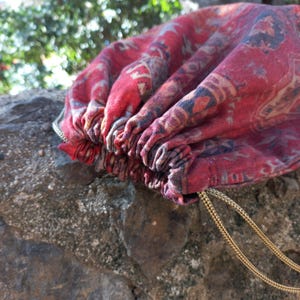May include: A red and blue patterned drawstring bag with gold cords. The bag is made of fabric and has a gathered top.