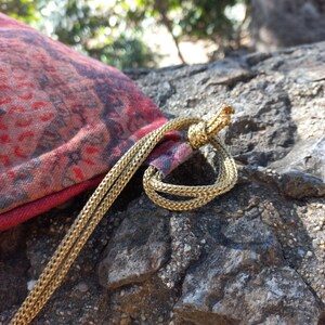 May include: A red and black patterned drawstring bag with a gold cord. The bag is lying on a rocky surface.