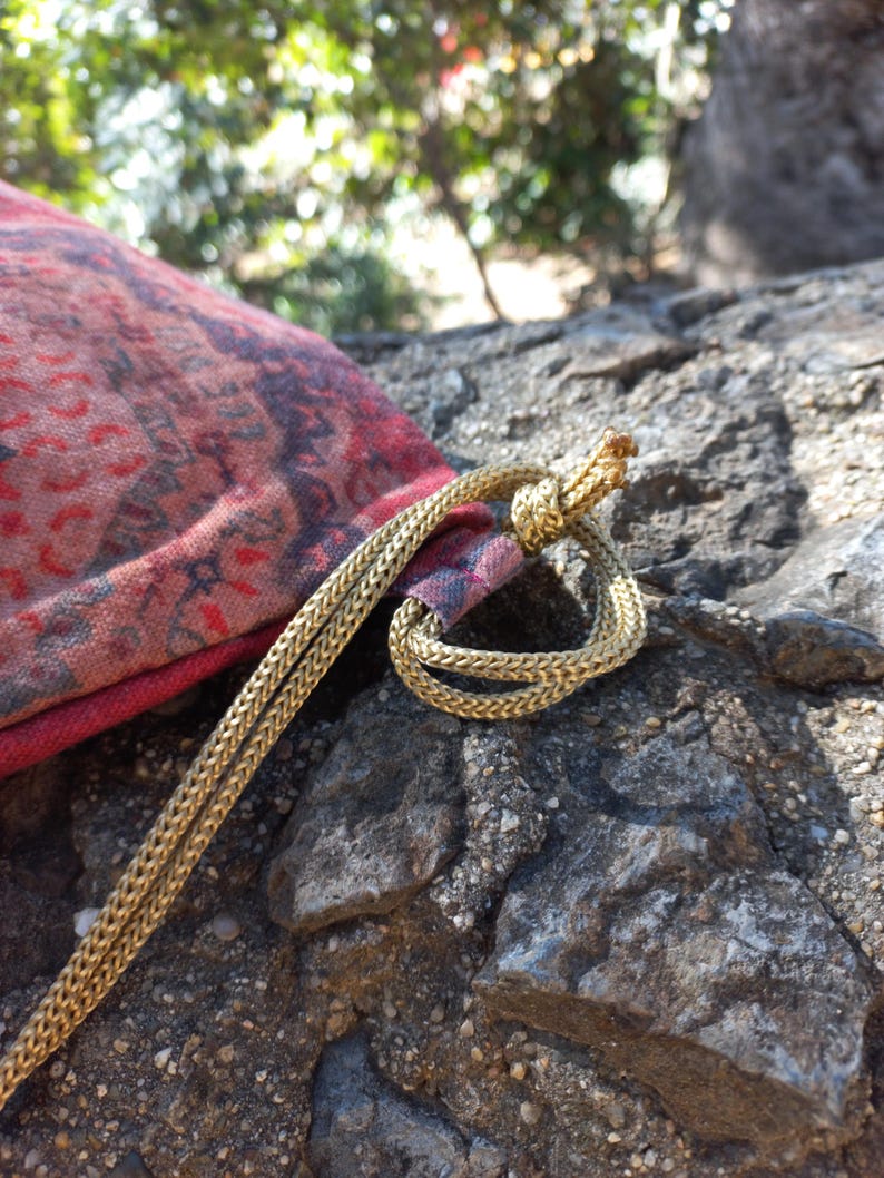 May include: A red and black patterned drawstring bag with a gold cord. The bag is lying on a rocky surface.