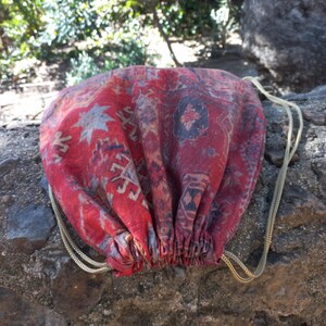 May include: A red and blue patterned drawstring bag with gold cords. The bag is made from a fabric that resembles a rug.
