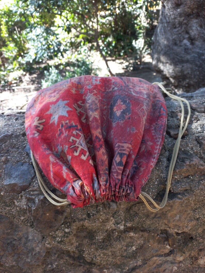 May include: A red and blue patterned drawstring bag with gold cords. The bag is made from a fabric that resembles a rug.