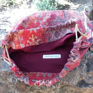 May include: Red drawstring bag with a patterned fabric design. The bag is lined with a burgundy fabric and has a white label that reads "Tilda & Matilda".