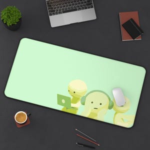 May include: A light green rectangular desk mat featuring a cartoon design of three figures with laptops and headphones. A white mouse is on the right side of the mat. A small potted plant, a laptop, and a coffee cup are also visible.