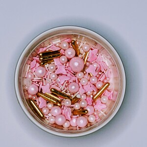 Peut inclure: Un bol blanc rempli de vermicelles roses et dorés. Les vermicelles comprennent de petites étoiles, des cœurs et des vermicelles ronds, ainsi que des vermicelles plus gros de type perle et des bâtonnets dorés.