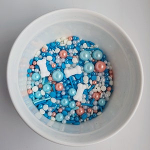 Könnte beinhalten: Eine weiße Schüssel gefüllt mit blauen, weißen und pfirsichfarbenen Streuseln. Die Streusel haben die Form von kleinen Kugeln, langen dünnen Stäbchen und Knochen.