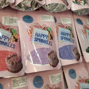 Può includere: Assortimento di piccoli sacchetti di codette colorate, etichettati "Happy Sprinkles". Le codette sono di vari colori, tra cui rosa, viola, blu e nero. Ogni sacchetto ha una piccola immagine di una torta con codette sopra.