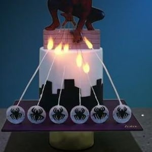 Op de afbeelding: Een taart met twee lagen met een Spider-Man-figuur erop. De taart heeft een witte basis met een zwart stadsgezichtontwerp. Zes spinnenwebachtige draden strekken zich uit vanaf de taart, elk eindigend in een bol met een spinnenembleem. Aangestoken kaarsen zijn aan de taart bevestigd.
