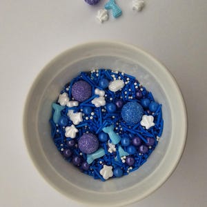 Può includere: Una ciotola bianca piena di zuccherini blu e viola, tra cui code di sirena, conchiglie e piccole sfere. Ci sono anche alcuni zuccherini sparsi sulla superficie bianca intorno alla ciotola.