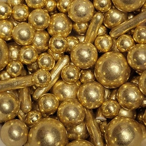 Könnte beinhalten: Nahaufnahme einer Sammlung goldfarbener essbarer Streusel. Die Streusel umfassen runde Kugeln unterschiedlicher Größe und längliche Stabformen. Die Streusel haben ein metallisches, glänzendes Finish, was darauf hindeutet, dass sie zum Dekorieren von Backwaren bestimmt sind.