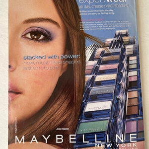 May include: A close-up of a woman's eye with eyeshadow applied. The image features a variety of eyeshadow palettes in blue, green, and purple shades. The text "new expert wear all day, crease-proof shadow" is featured at the top of the image. The text "MAYBELLINE NEW YORK MAYBE SHE'S BORN WITH IT. MAYBE IT'S MAYBELLINE." is featured at the bottom of the image.