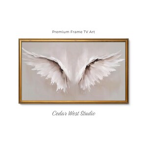 May include: A rectangular wall art piece featuring a pair of white feathered wings painted in a soft, ethereal style against a neutral background, enclosed in a decorative gold frame. The text Premium Frame TV Art and Cedar West Studio appear.