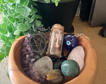 May include: A round wooden bowl containing a collection of items including a small glass jar with a cork stopper, a black pentagram charm, a blue glass evil eye bead, a strand of amethyst beads, and several polished tumbled gemstones.